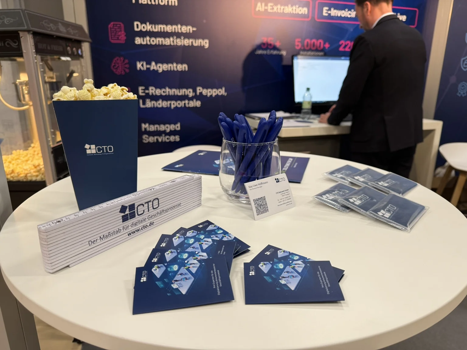 Stehtisch am CTO-Messestand mit Popcorn im CTO-Becher, Kugelschreibern, einem Zollstock mit dem Slogan „Der Maßstab für digitale Geschäftsprozesse“ und Infoflyern.